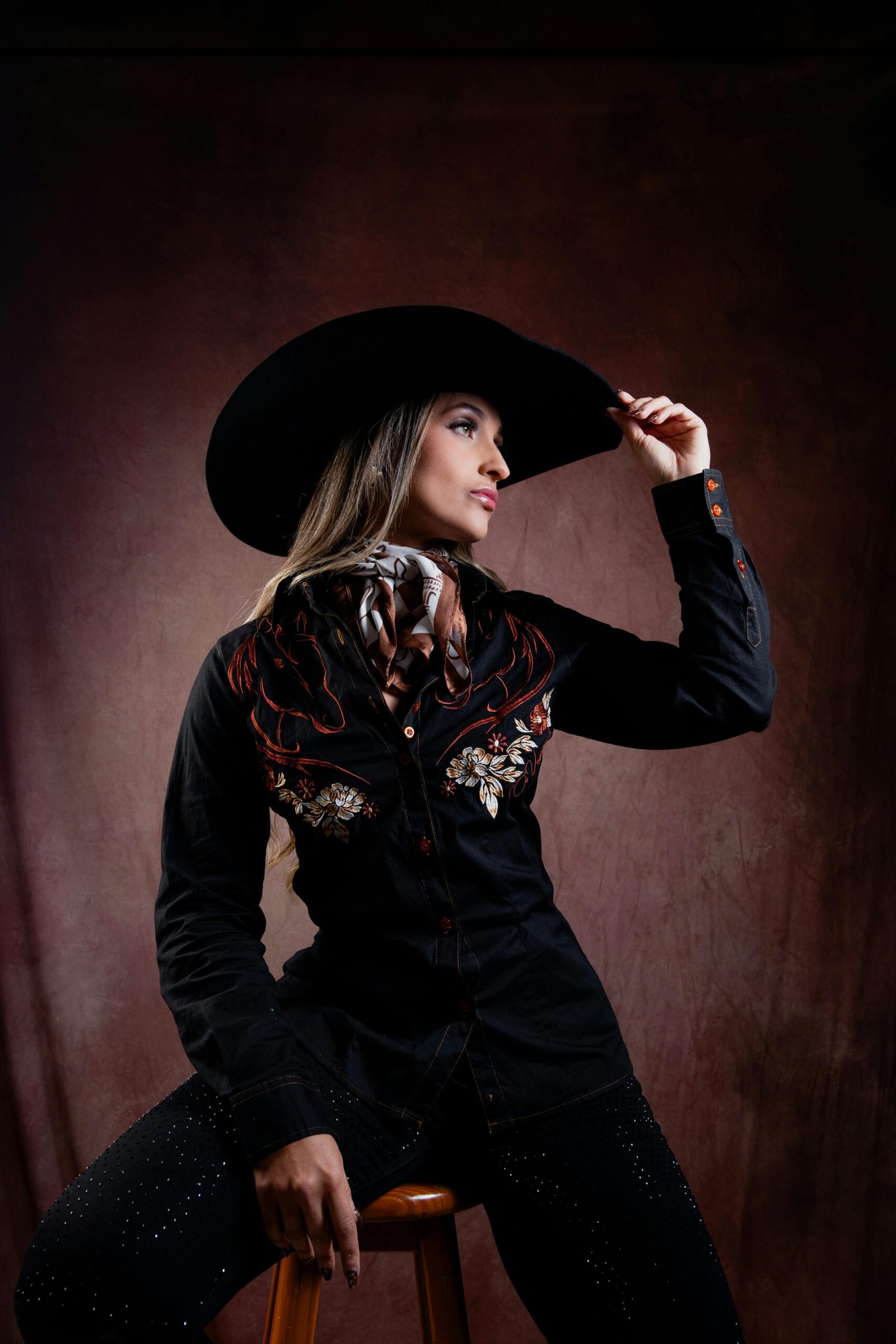 Stylish woman in western attire and cowboy hat on a stool, exuding confidence.