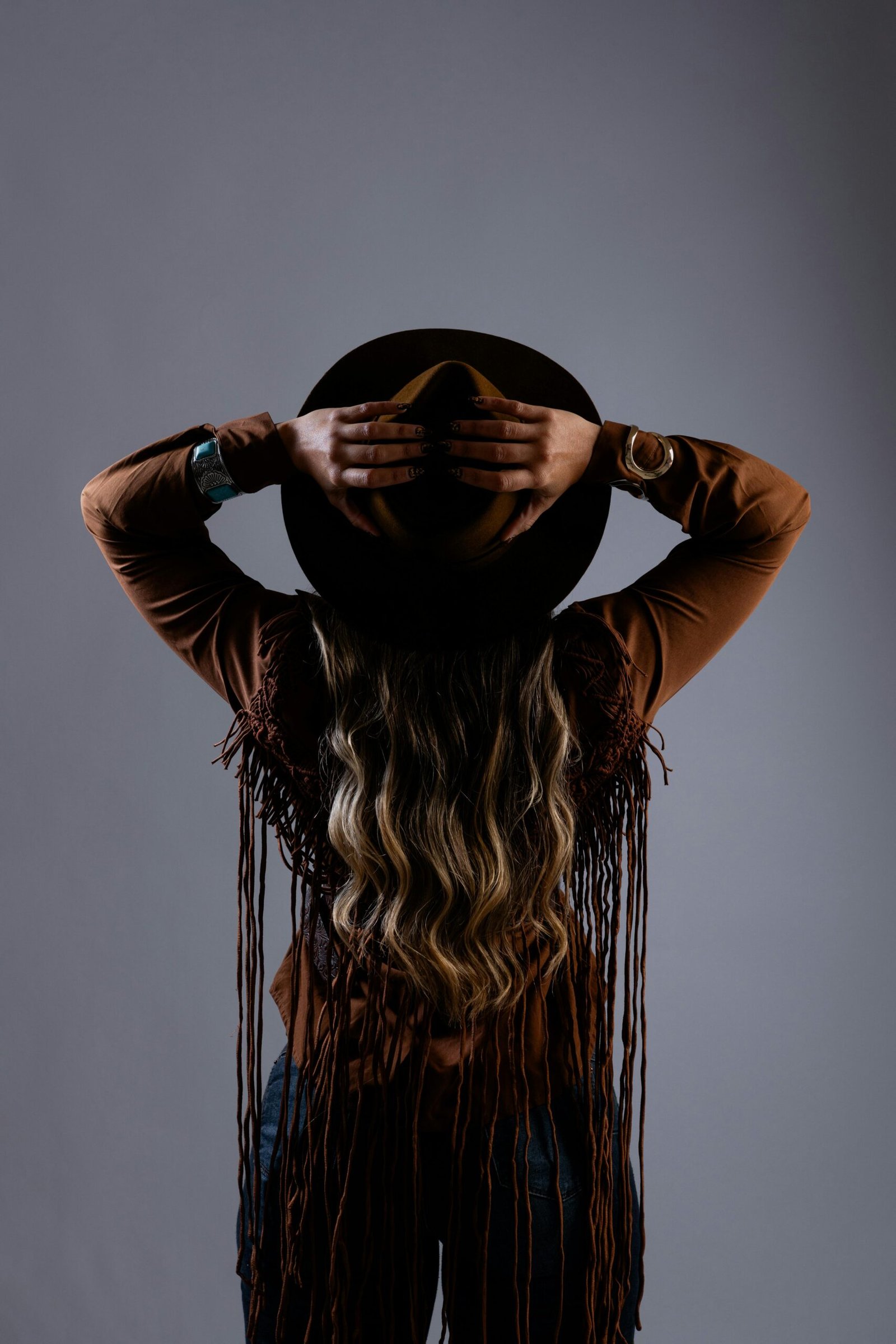 Woman in a brown cowboy hat and fringed jacket seen from behind in studio lighting.